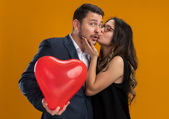 男朋友一对幸福美丽的情侣用心形的红气球亲吻她的男朋友 在橙色的墙上庆祝情人节心礼物吻