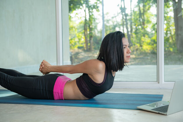 亚洲运动型女子练习瑜伽 背部和臀部训练姿势 在笔记本电脑上观看在线健身视频教程 在家里坐在客厅的垫子上练习互联网老师健康