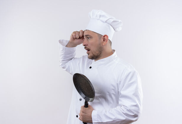 头部印象深刻的年轻帅哥厨师穿着厨师制服拿着煎锅站在侧视图看一边手放在头上隔离在白色的墙上帅气年轻制服