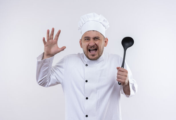 愤怒愤怒的年轻帅哥厨师身着厨师制服 手举勺子 站在孤零零的白墙上手持勺子厨师