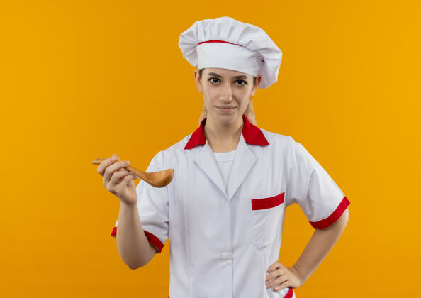 漂亮自信的年轻漂亮厨师身着厨师制服 手拿勺子放在腰上 隔离在橙色的墙上手腰制服