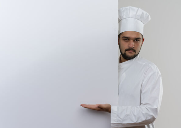 制服身着厨师制服的自信年轻男厨师站在身后 用手指着隔离在白色墙壁上的白色墙壁 并留有复印空间墙厨师指向