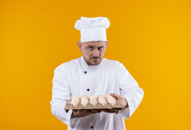 厨师自信的年轻帅哥厨师身着厨师制服 手里拿着一盒鸡蛋 隔离在橙色的墙上制服厨艺纸箱