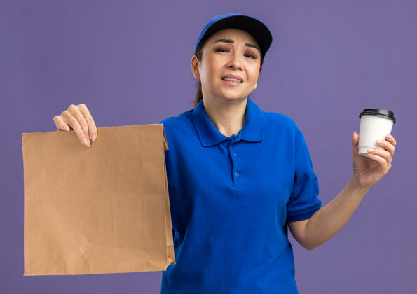 女人身穿蓝色制服 戴着帽子 手里拿着纸包和纸杯的年轻女送货员站在紫色的墙上 对悲伤的表情感到恼火杯子站着包装