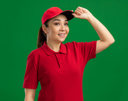 年轻身穿红色制服 戴着帽子的年轻女送货员微笑着 自信地抚摸着站在绿墙上的帽子女人帽子自信