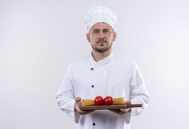 烹饪自信的年轻帅哥厨师身着厨师制服 手拿着砧板 上面有西红柿和意大利面 隔离在白色的墙上年轻持有意大利面
