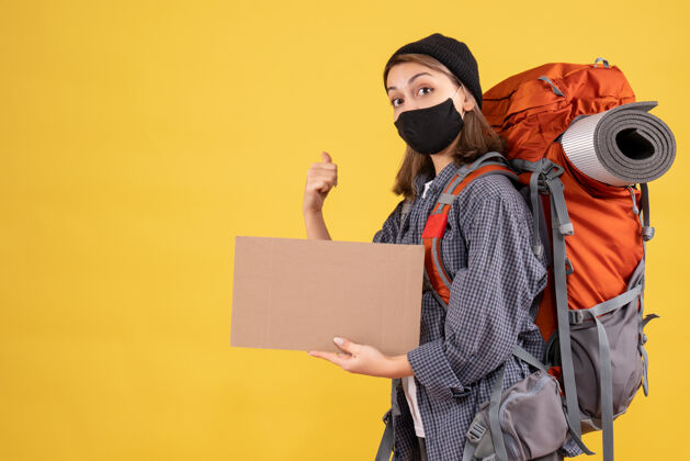 纸板带着黑色面具和背包的旅行女孩拿着纸板指着后面的黄色成人背包面具