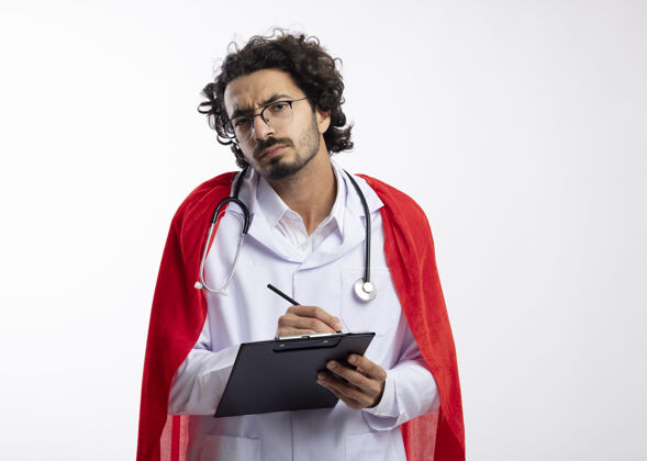 年轻无知的年轻白种人超级英雄戴着眼镜 身穿医生制服 披着红色斗篷 脖子上戴着听诊器 手里拿着铅笔和剪贴板男制服听诊器