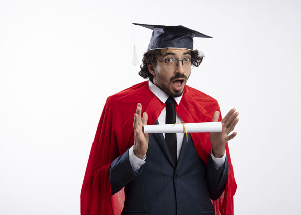 复制惊讶的年轻白种人超级英雄戴着眼镜 穿着红色斗篷的西装 戴着毕业帽 手里拿着毕业证书 看着镜头帽子惊喜持有