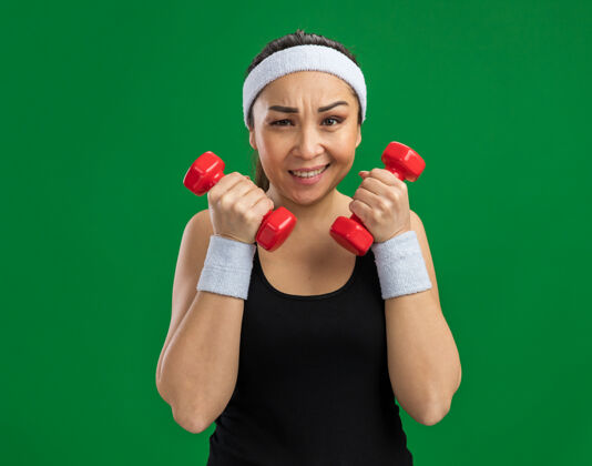 头带戴着头带和哑铃做运动的年轻健身女士紧张而自信地站在绿色的墙上女人锻炼紧张