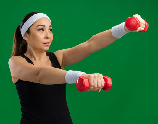 锻炼戴着头带和哑铃做运动的年轻健身女士紧张而自信地站在绿色的墙上紧张年轻健身