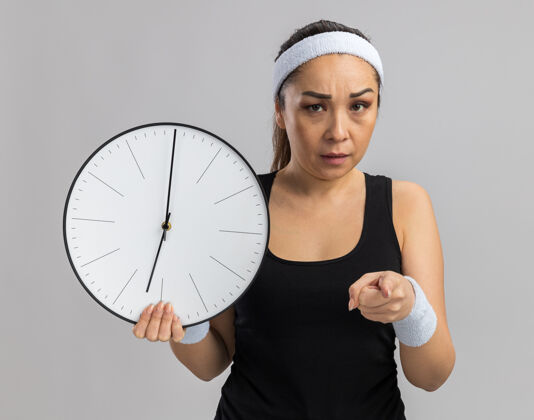 女人戴着头带和臂章拿着挂钟用食指指着的年轻健身女士站在白色的墙上生气了目录手指健身