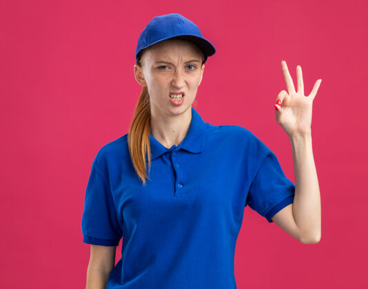 女孩身穿蓝色制服 头戴鸭舌帽的年轻女送货员不高兴地站在粉红色的墙上 显示“ok”标志站着制服展示