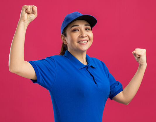 年轻身穿蓝色制服 头戴鸭舌帽的年轻女送货员站在粉红色的墙上 高兴而兴奋地握紧拳头握紧制服兴奋