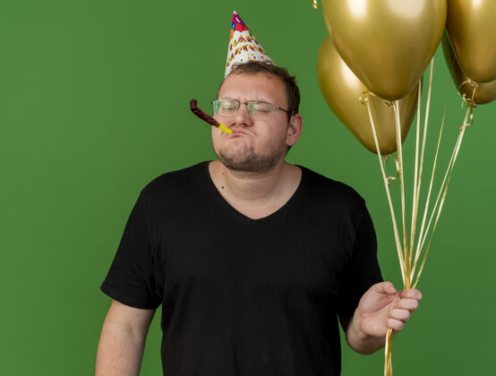 聚会戴着生日帽戴着光学眼镜的未被释放的成年斯拉夫男子手持氦气球吹派对哨子帽子举行生日
