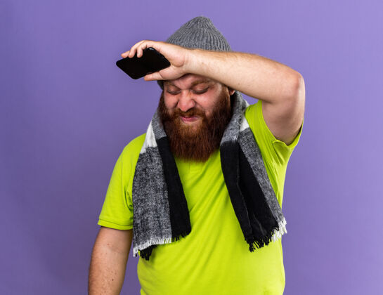 流感戴着帽子 脖子上围着暖和的围巾 留着不健康的胡须 感觉很可怕 患了流感 拿着手机 站在紫色的墙壁上很失望电话感觉温暖