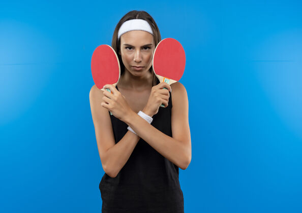 腕带自信的年轻漂亮的运动女孩戴着头带和腕带拿着乒乓球拍隔离在蓝色的墙壁与复制空间乒乓球头带漂亮