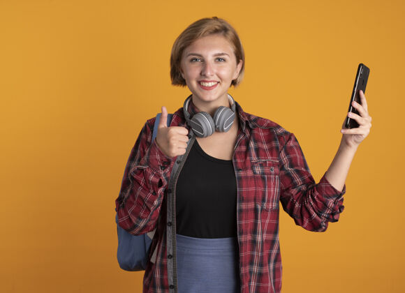 女孩微笑着的年轻斯拉夫女学生戴着耳机 背着背包 竖起大拇指 拿着电话年轻微笑背包