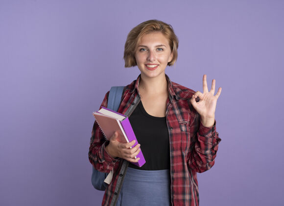 女孩微笑的年轻斯拉夫学生女孩背着书包拿着书和笔记本手势ok手势标志学生穿着