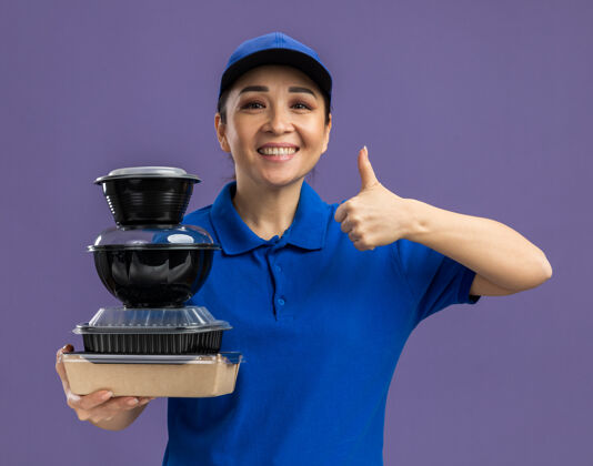 微笑身着蓝色制服 头戴鸭舌帽 手持一叠食品包装的快乐年轻女送货员站在紫色的墙上 愉快地微笑着竖起大拇指帽子食物女人