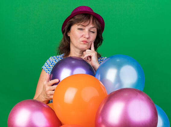 表情中年妇女戴着派对帽 带着五颜六色的气球 带着怀疑的表情站在绿色的墙上庆祝生日派对中间女人怀疑