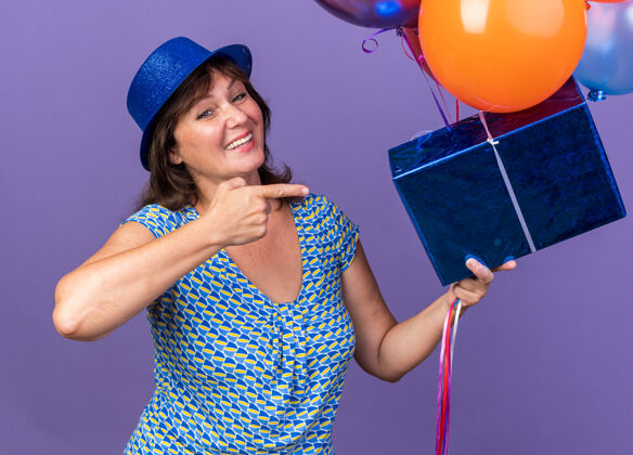 五颜六色快乐快乐的中年妇女戴着派对帽 手里拿着一束五颜六色的气球和礼物 用食指指着它微笑着站在紫色的墙上庆祝生日派对庆祝年龄微笑