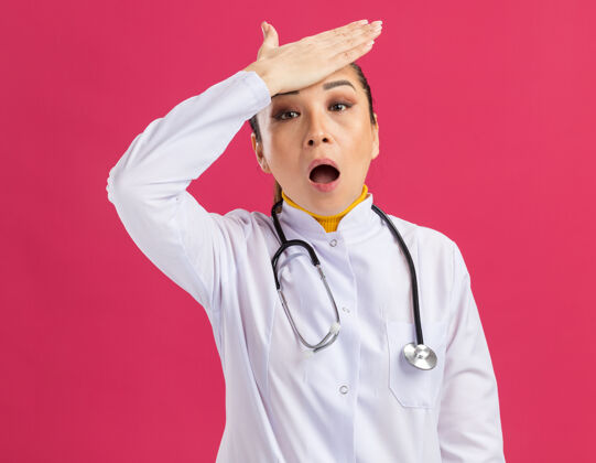 额头年轻的女医生 穿着白药衣 脖子上戴着听诊器 额头上戴着一只手 误站在粉红色的墙上手女人困惑