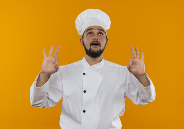 年轻令人印象深刻的年轻男厨师在厨师制服抬头看了看 并做了好的迹象隔离在橙色的墙上厨师好的印象深刻