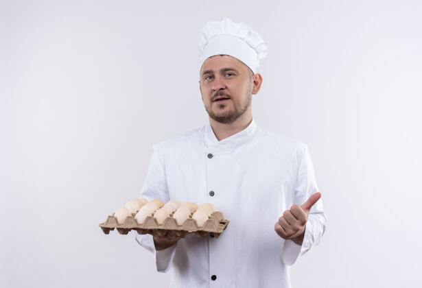 年轻自信的年轻帅哥厨师穿着厨师制服 拿着一盒鸡蛋 在白色的墙上孤立地竖起大拇指鸡蛋烹饪大拇指