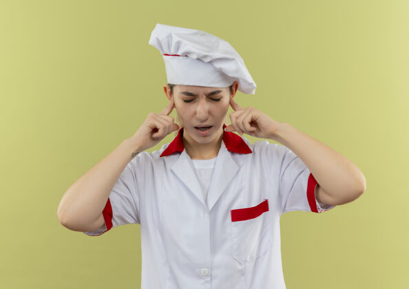制服年轻漂亮的厨师穿着厨师制服 把手指放在耳朵里 闭着眼睛孤立在绿色的墙上恼火手指厨师