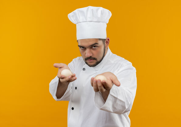 制服自信的年轻男性厨师身着厨师制服 将鸡蛋伸向橙色墙壁上的隔离空间走向烹饪伸展