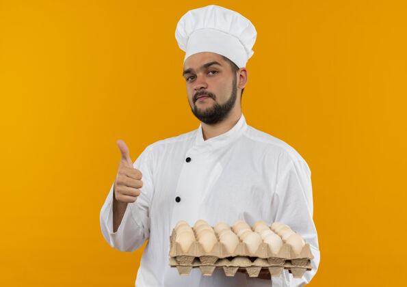 制服自信的年轻男性厨师身着厨师制服 手拿一盒鸡蛋 在橙色的墙上孤立地竖起大拇指信心年轻持有