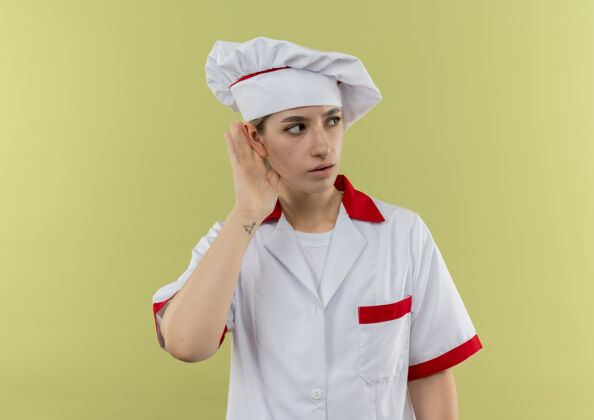 制服好奇的年轻漂亮厨师穿着厨师服做着听不到你的手势看着隔离在绿墙上的一面厨师壁板手势