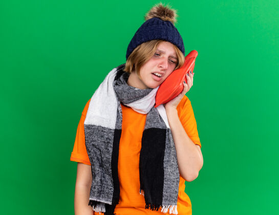 温暖不健康的年轻女子穿着橙色t恤 戴着帽子 围着暖和的围巾 站在绿色的墙上拿着一个患感冒和流感的热水瓶 感觉很糟糕抱着不健康瓶子