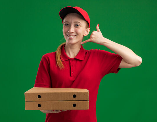 制造身穿红色制服 头戴鸭舌帽 拿着披萨盒 做着“叫我”手势 微笑自信地站在绿色墙壁上的年轻送货女孩微笑站着披萨