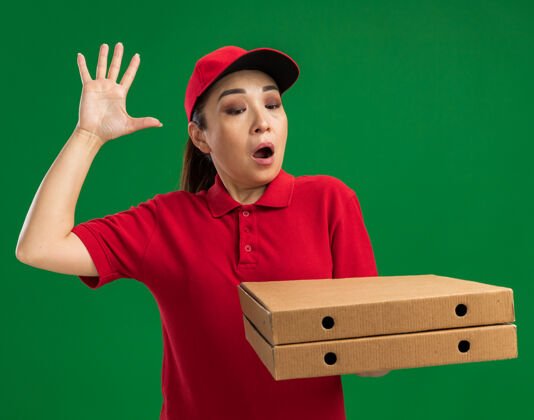 盒子年轻的送货员身穿红色制服 头戴鸭舌帽 手举披萨盒 站在绿色的墙上 看上去既惊讶又惊讶惊喜女人抱着