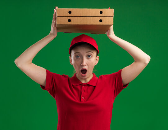 女孩身穿红色制服 头戴鸭舌帽 头顶披萨盒的年轻送货员站在绿色的墙壁上 惊讶不已披萨制服人头