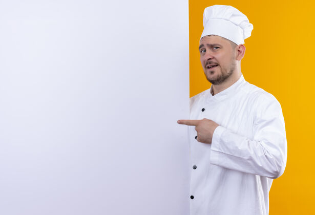站立穿着厨师制服的年轻帅哥站在白色的墙后 指着隔离在橙色墙上的复制空间烹饪墙壁制服