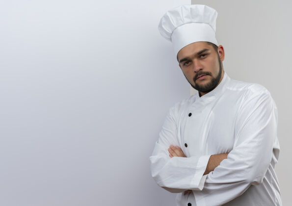 自信自信的年轻男性厨师身着厨师制服站在白色墙壁前 封闭的姿势看起来孤立在白色墙壁与复制空间前面年轻封闭