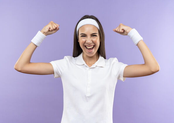 运动快乐漂亮的运动女孩戴着头带和腕带在紫色的墙上做着强烈的孤立的手势穿着漂亮快乐