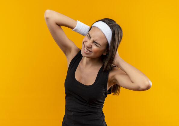 运动疼痛的年轻漂亮的运动女孩戴着头带和腕带把双手放在她的头后面橙色的墙上戴疼头带
