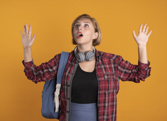 年轻人震惊的年轻斯拉夫女学生戴着耳机背着背包站着举起手空间橙色学生