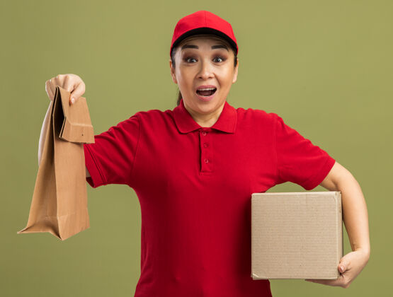 女人身穿红色制服 头戴鸭舌帽 手拿纸包和纸箱的年轻送货员站在绿色的墙边 又高兴又惊讶帽子惊喜年轻