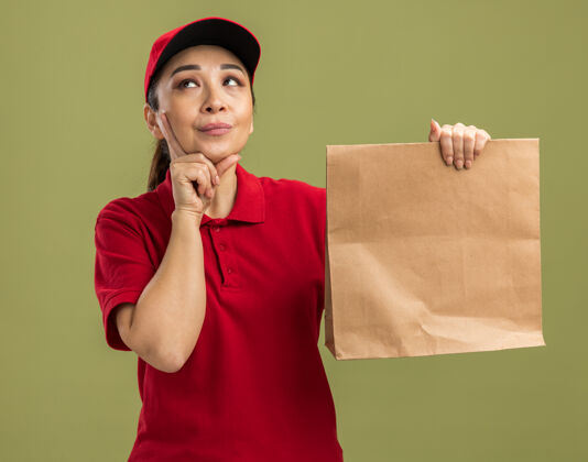 站着年轻的送货员身穿红色制服 戴着帽子 手里拿着纸包 站在绿色的墙上 困惑地抬头望着女人年轻帽子