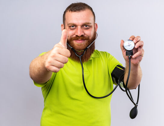 不健康不健康的胡子男人穿着黄色马球衫感觉更好用血压计测量血压微笑着竖起大拇指站在白色的墙上压力拇指测量