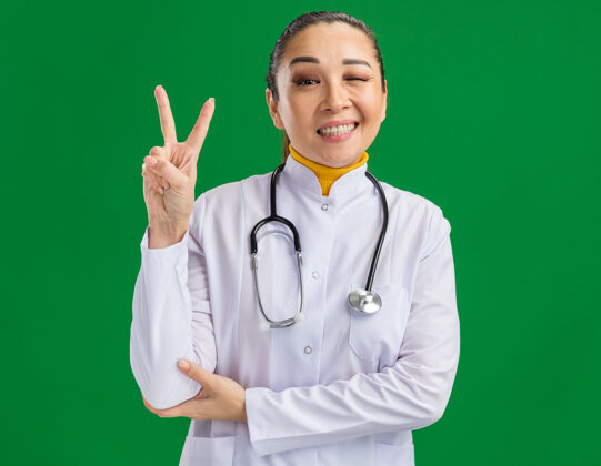 快乐身穿白色药衣 脖子上戴着听诊器的年轻女医生快乐地眨着眼睛 微笑着 绿色的墙上立着v字标志医生手势表演