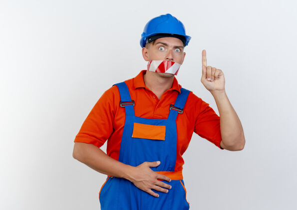 头盔年轻的男建筑工人穿着制服 头戴安全帽 惊讶地用胶带封住了嘴 指着他制服嘴胶带