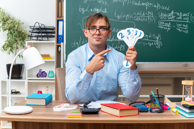 坐着年轻的男老师戴着眼镜 展示着车牌 在教室的黑板前微笑着看着书桌上的书本和笔记数学男老师