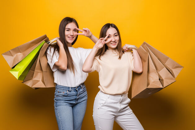 快乐兴奋的年轻女性女孩朋友们拿着购物包装袋在黄色的墙上摆着孤立的姿势购物人们的生活方式理念正面女性购买