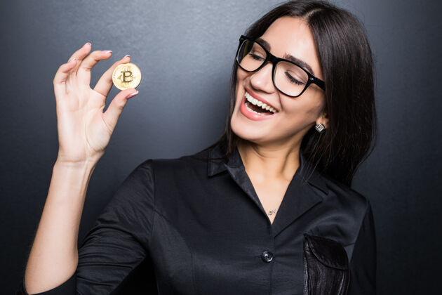 企业家戴着眼镜的年轻成功自信女人手里拿着一枚比特币 孤零零地贴在黑墙上金融加密比特币